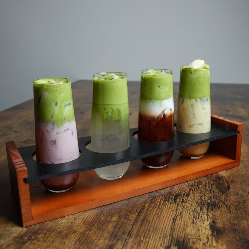 Four colorful layered Thai drinks in a wooden flight holder on a rustic wooden table.