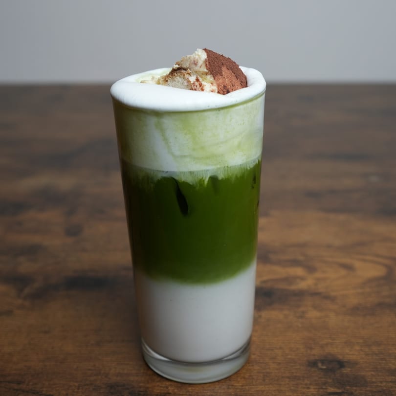 Nalak Thai's layered iced matcha latte with whipped cream and cocoa powder on a wooden table.
